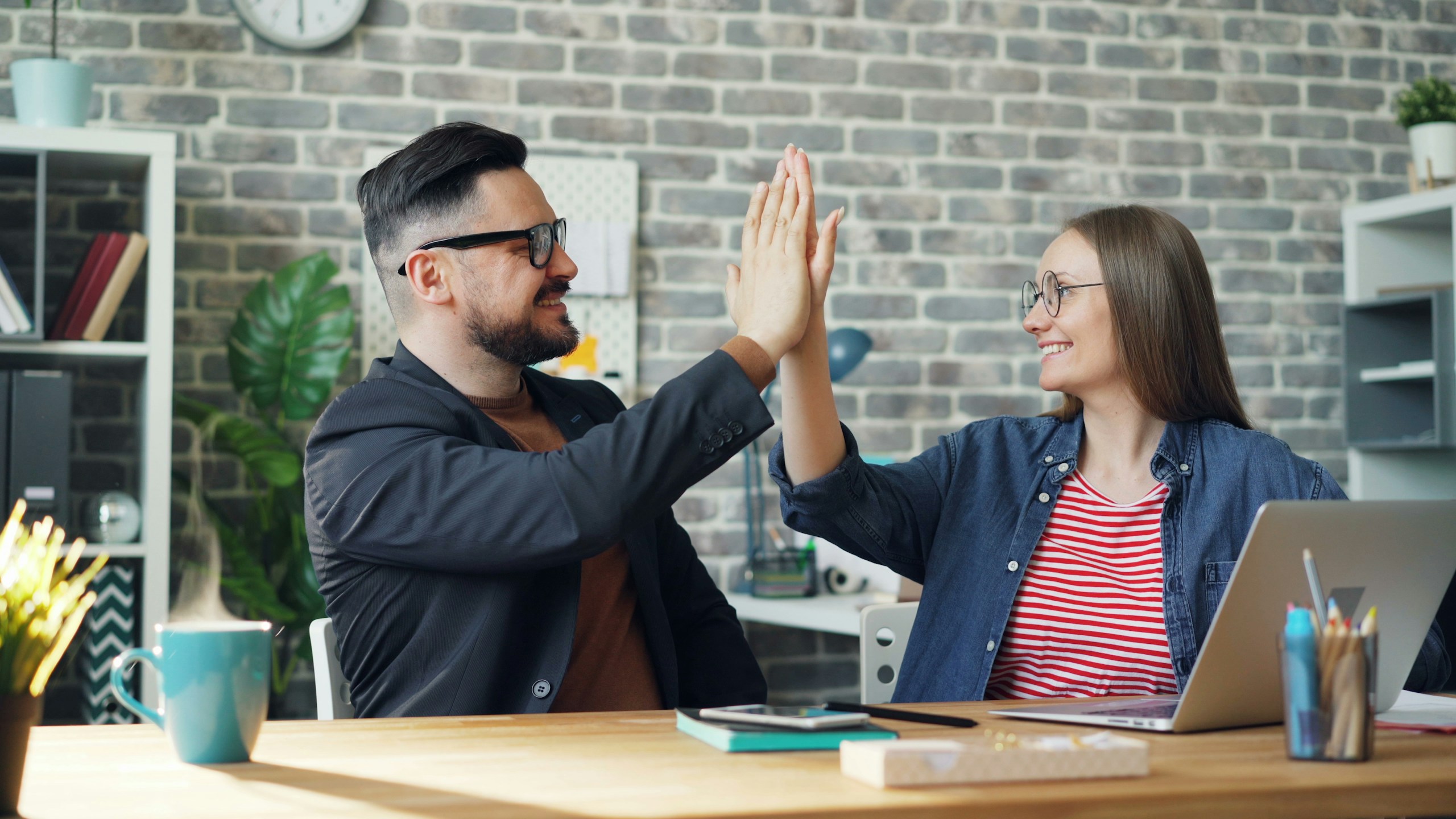 happy colleagues man and woman doing high-five sitting at table in office while cup of hot tea is steaming on desk. People and work concept.