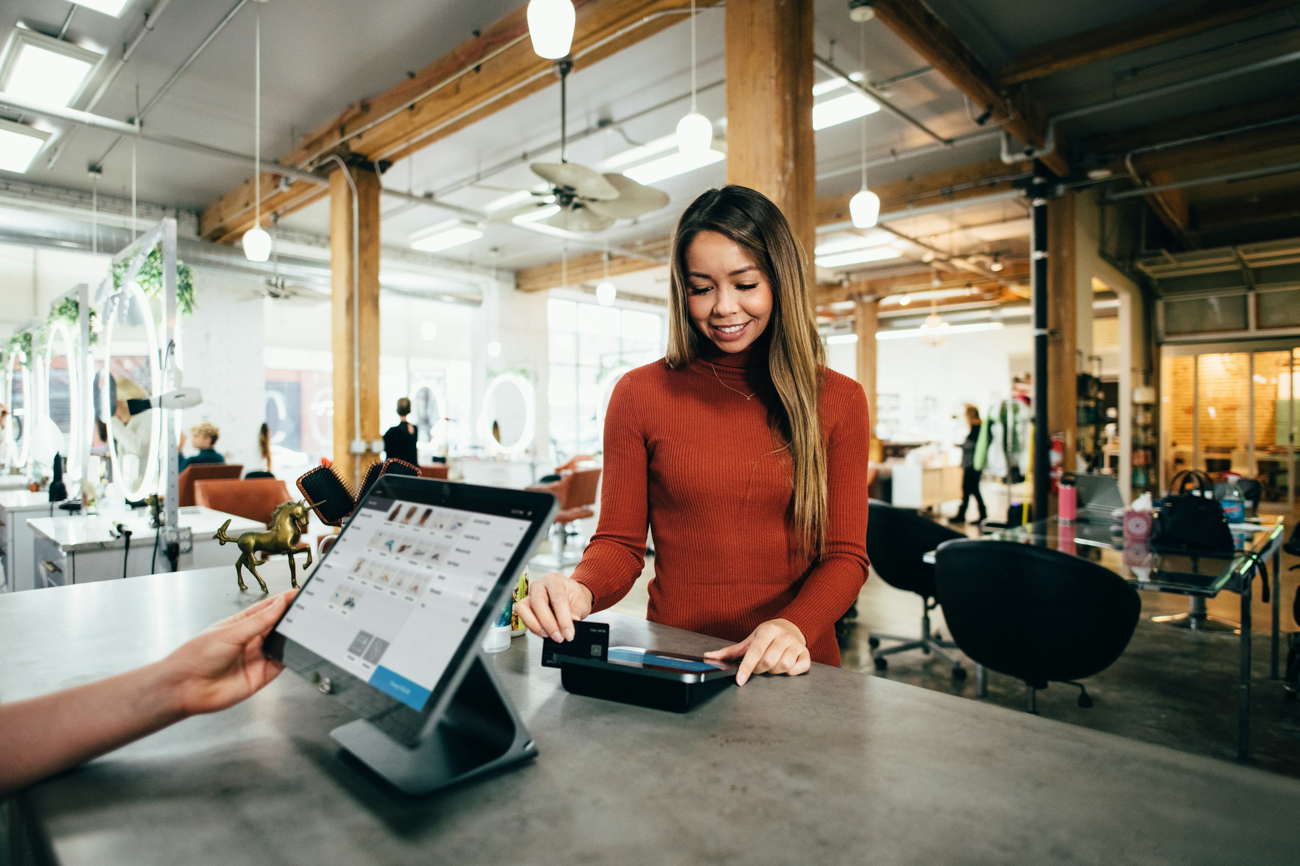 Smiling woman makes contactless payment at modern retail POS terminal with tablet register system. Industrial-style store interior showcases contemporary cashless shopping experience.
