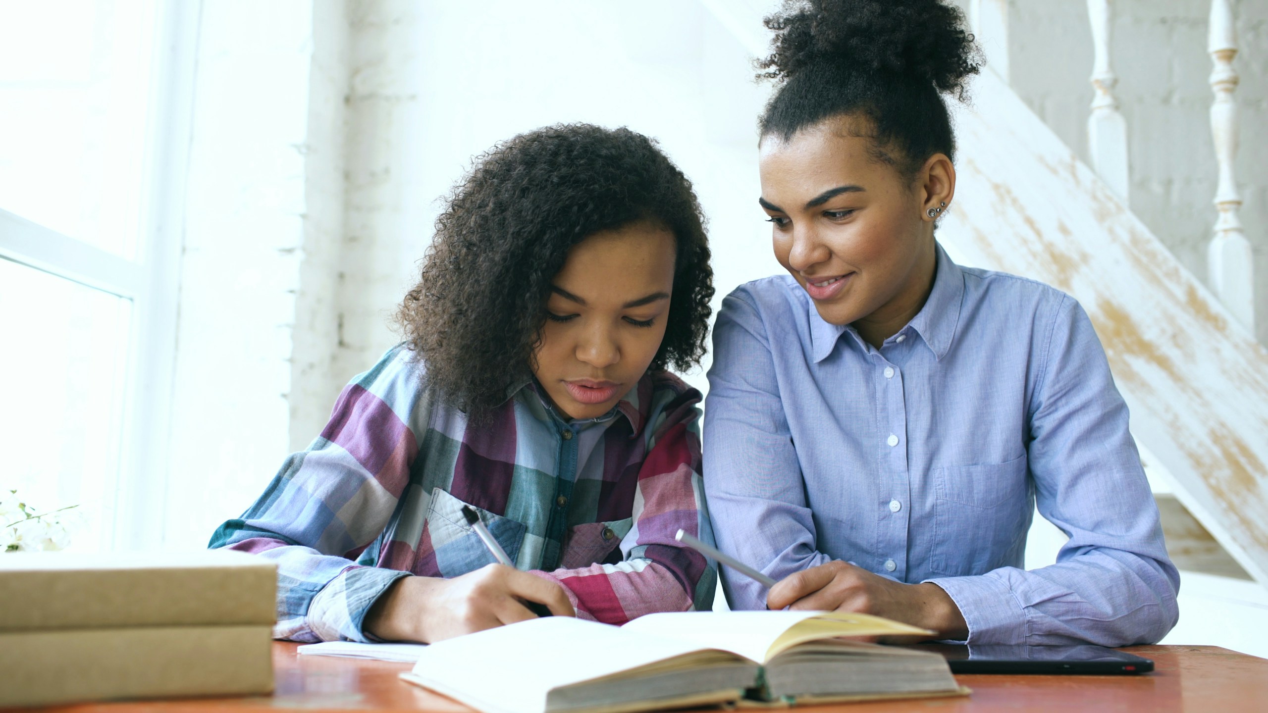 Teenage curly haired mixed race young girl sitting at the table concentrating focused learning lessons and her elder sister helps her studying at home